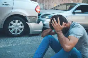 Man in pain and sad while sitting on ground with car accident scene in background